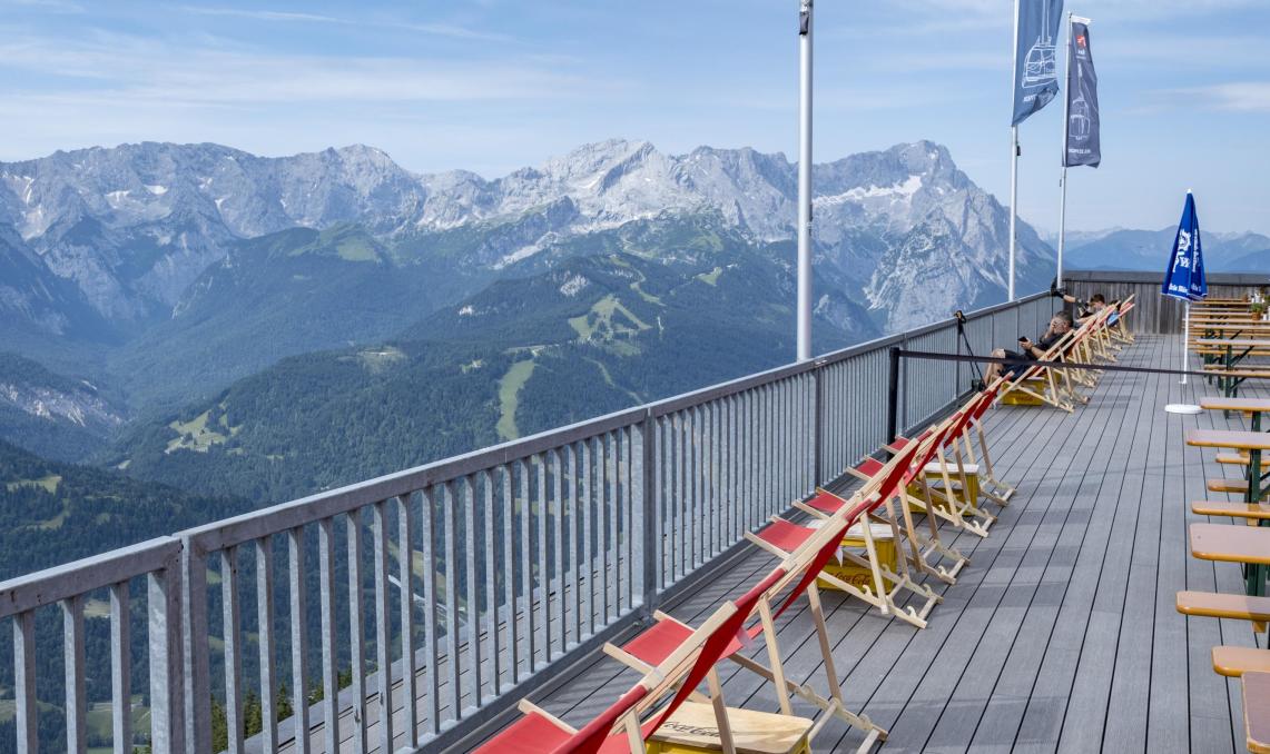 Terrasse panoramique en bois composite foncé avec garde-corps métallique, transats en bois avec toiles rouges alignés face à une vue spectaculaire sur les montagnes, drapeaux flottant au vent, point d'observation en altitude